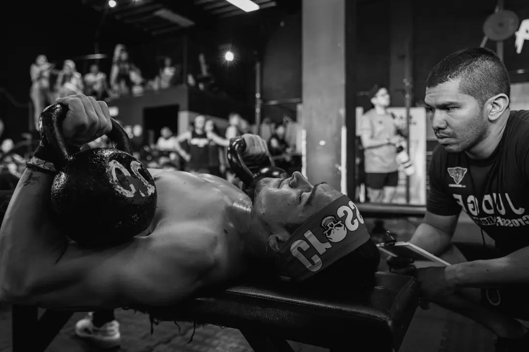 A judge watching an athlete hold two kettlebells on a bench, in a crowded competition gym